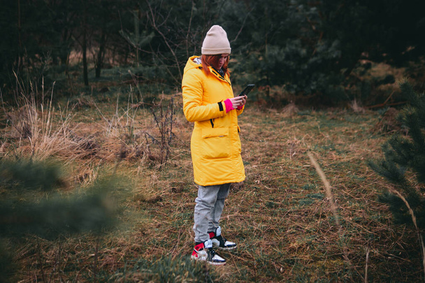 Una niña, una mujer con una chaqueta larga de invierno amarillo se encuentra en medio de la naturaleza salvaje, el bosque. Mira su smartphone. Concepto de navegación, comunicación. Piérdete. - Foto, imagen
