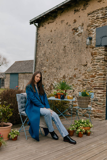 Foto de larga duración de la chica caucásica bonita con el pelo suelto con abrigo azul, jeans y botas sentados en la terraza de verano con flores y descanso en el día de fin de semana de primavera.  - Foto, imagen