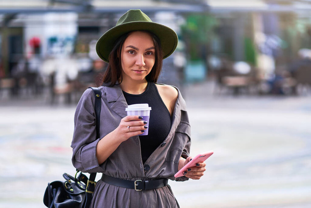 Woman traveler wearing felt hat and overalls with a backpack holds paper cup. Female drinking coffee and using smartphone during walking in the center of a european city - Photo, image