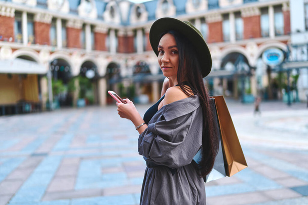Stylish young cute attractive hipster woman shopaholic wearing felt hat with paper shopping bags using phone and walks in the center of a european city during black friday sales week  - Fotografie, Obrázek