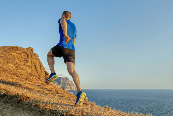 back male runner run uphill mountain trail in blue sky background, summer jogging along coast at sunset - Photo, Image