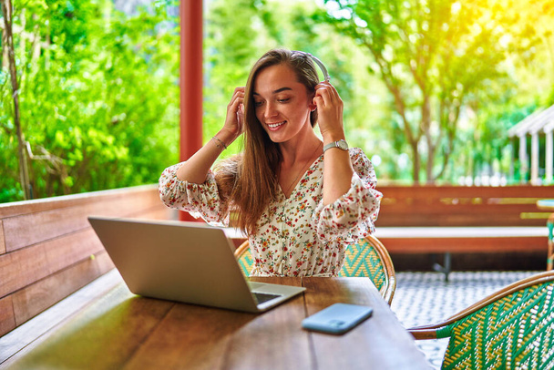 Happy cheerful smiling beautiful cute joyful young millennial girl wearing wireless headphones using laptop for remote work, social networking and online browsing at green cafe  - Foto, afbeelding