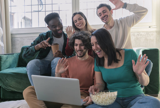 Kanepede oturmuş bilgisayar ekranındaki el sallamalarına bakıp gülen beş çok ırklı arkadaş eşle uzaktan iletişimin keyfini çıkarıyorlar. Video çağrısı yap, webcam modern teknoloji konseptiyle internetten selamla - Fotoğraf, Görsel