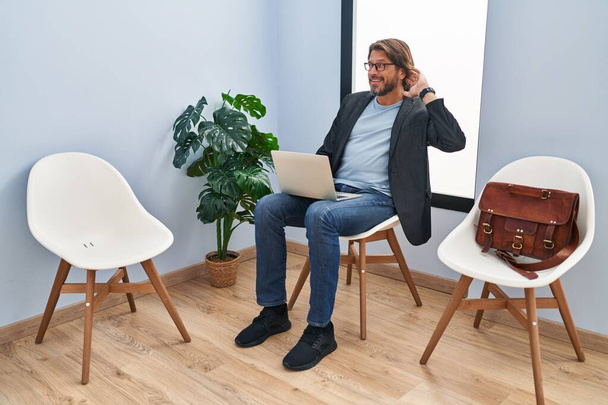 Un bel homme d'âge moyen assis dans une salle d'attente travaillant avec un ordinateur portable souriant, la main sur l'oreille, écoutant une rumeur ou des commérages. concept de surdité.  - Photo, image