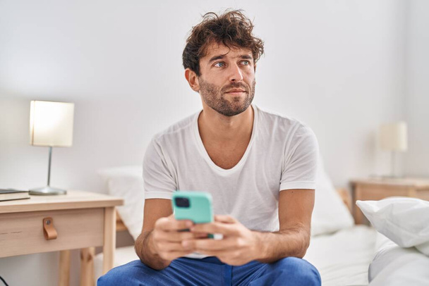 Молодий чоловік використовує смартфон з серйозним виразом, сидячи в ліжку в спальні
 - Фото, зображення