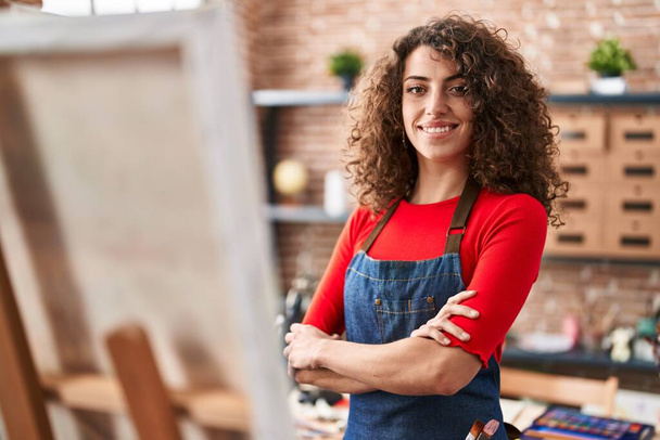 Genç İspanyol kadın sanatçı gülümsüyor. Kollarını kavuşturup sanat stüdyosunda duruyor. - Fotoğraf, Görsel