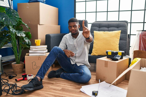 Hombre afroamericano sentado en el suelo en una casa nueva señalando con el dedo hacia arriba y la expresión enojada, sin mostrar ningún gesto  - Foto, imagen