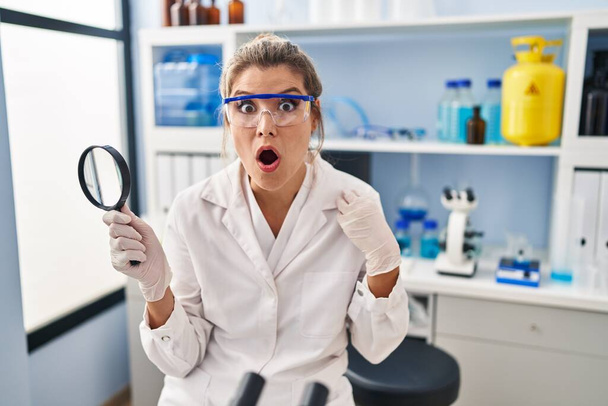 Jovem mulher trabalhando no laboratório cientista segurando lupa assustada e espantada com a boca aberta para surpresa, rosto descrença  - Foto, Imagem