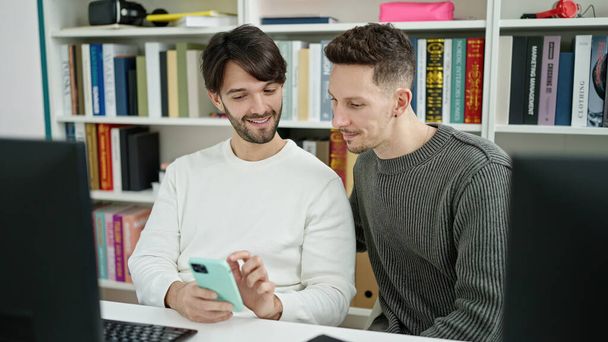 Két férfi diák számítógépet és okostelefont használ a könyvtár egyetemen. - Fotó, kép