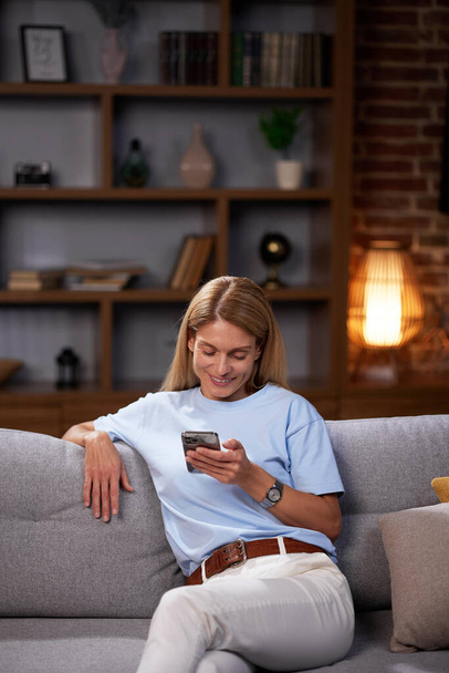 Chica rubia sentada en el sofá mira en el teléfono inteligente, charlando, relajarse en casa. Sonriente mujer adulta se comunica amigable escribiendo un mensaje en el teléfono - Foto, Imagen