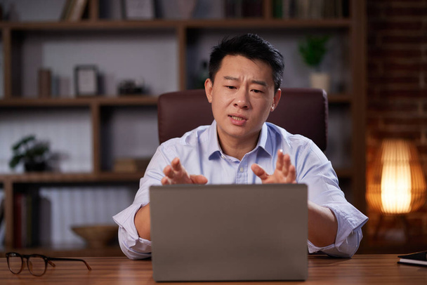 Asian man holding online webinar using laptop webcam, sitting in a home office. Man having online meeting and communicating remotely, looking at computer screen. - Foto, Bild