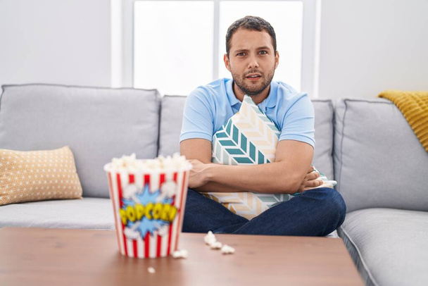 Hombre hispano viendo televisión en casa sentado en el sofá expresión despistada y confusa. concepto de duda.  - Foto, Imagen