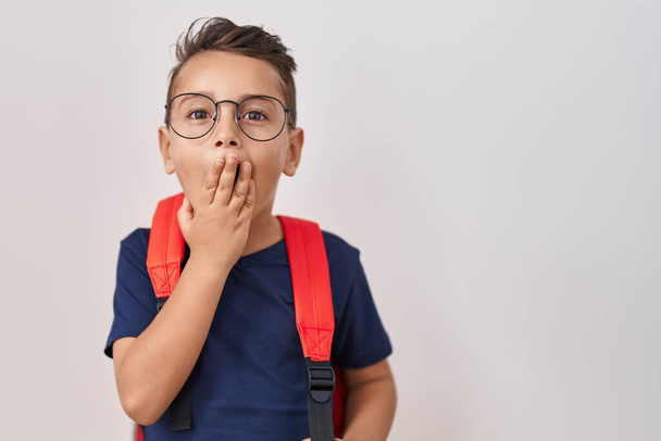 小さなヒスパニック系の少年が眼鏡をかけ、学生のバックパックを手で口をカバーし、ショックを受け、ミスを恐れている。驚きの表情  - 写真・画像