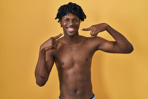 Jonge Afrikaanse man met dreadlocks staat shirtloos lachend vrolijk te tonen en te wijzen met vingers tanden en mond. tandheelkundige gezondheidszorg.  - Foto, afbeelding