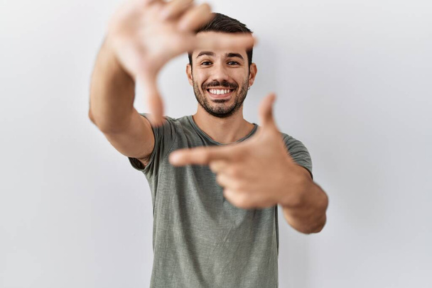Jonge Spaanse man met baard draagt casual t-shirt over witte achtergrond glimlachende maken frame met handen en vingers met gelukkig gezicht. creativiteit en fotografie.  - Foto, afbeelding
