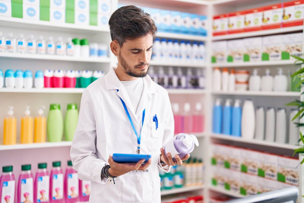 Joven farmacéutico hispano usando touchpad sosteniendo la botella de gel en la farmacia - Foto, imagen