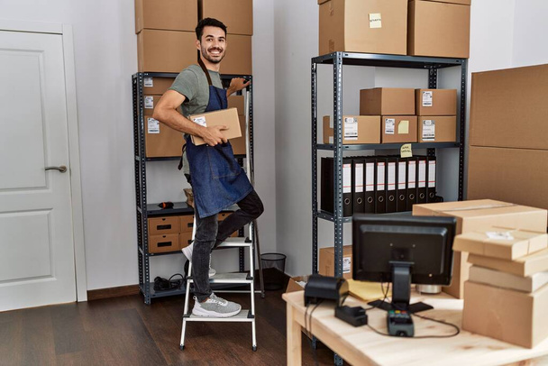 Young hispanic man business worker standing on ladder holding package at storehouse - Fotoğraf, Görsel