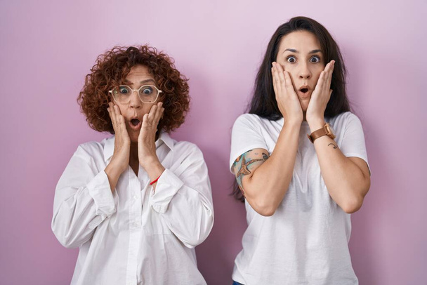 Hispanic mother and daughter wearing casual white t shirt over pink background afraid and shocked, surprise and amazed expression with hands on face  - Photo, image