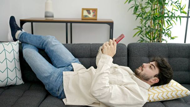 Νεαρός ισπανόφωνος άντρας που χρησιμοποιεί smartphone ξαπλωμένο στον καναπέ στο σπίτι - Φωτογραφία, εικόνα