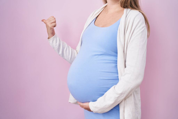 Young pregnant woman expecting a baby, touching pregnant belly pointing thumb up to the side smiling happy with open mouth  - Photo, Image