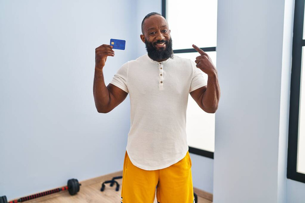 Africano americano vestindo sportswear segurando cartão de crédito sorrindo feliz apontando com mão e dedo  - Foto, Imagem