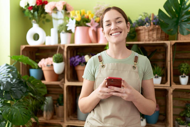 Junge schöne Floristin lächelt selbstbewusst mit Smartphone im Blumenladen - Foto, Bild