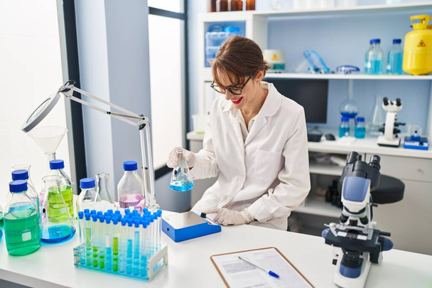 Young caucasian woman wearing scientist uniform measuring liquid at laboratory - 写真・画像