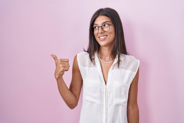 Morena jovem mulher de pé sobre fundo rosa usando óculos sorrindo com o rosto feliz olhando e apontando para o lado com o polegar para cima.  - Foto, Imagem