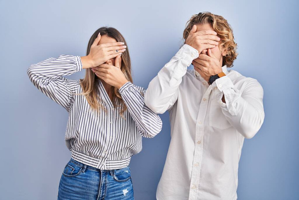 Young couple standing over blue background covering eyes and mouth with hands, surprised and shocked. hiding emotion  - Foto, Imagem