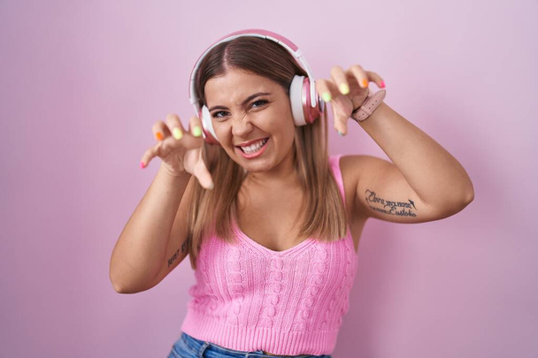 Giovane donna bionda che ascolta musica usando le cuffie sorridenti divertente facendo gesto artiglio come gatto, espressione aggressiva e sexy  - Foto, immagini