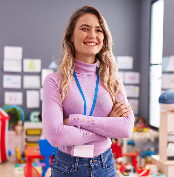 Young blonde woman teacher smiling confident standing with arms crossed gesture at kindergarten - Foto, afbeelding