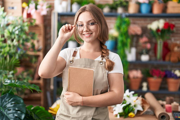 Young blonde woman florist smiling confident holding notebook at florist - Фото, изображение