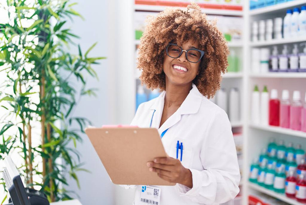 African american woman pharmacist smiling confident writing on document at pharmacy - Photo, Image