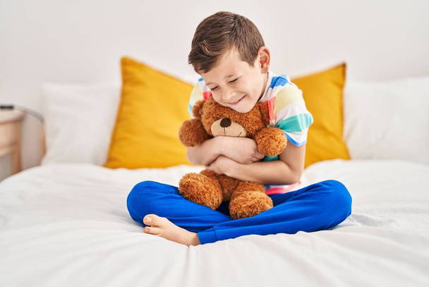 Blond child hugging teddy bear sitting on bed at bedroom - Foto, Imagen