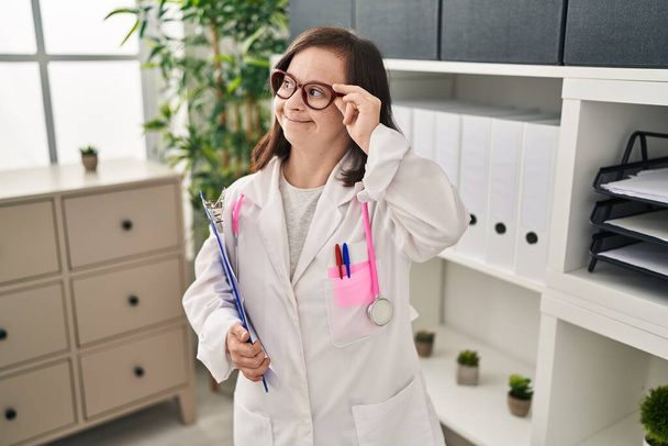Down syndrome woman wearing doctor uniform holding clipboard at clinic - Foto, Imagen