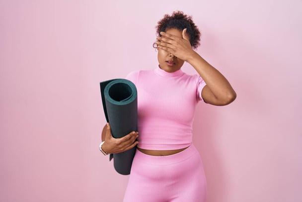 Junge hispanische Frau mit lockigem Haar, die eine Yogamatte über rosa Hintergrund hält und die Augen mit der Hand bedeckt, sieht ernst und traurig aus. Versteck- und Ablehnungskonzept  - Foto, Bild