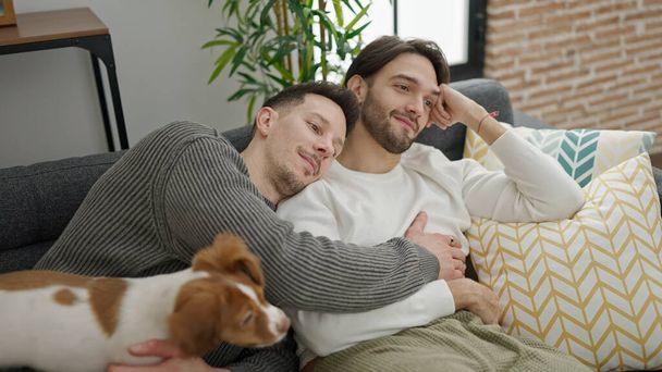 Двое мужчин обнимают друг друга сидя дома на диване с собакой - Фото, изображение