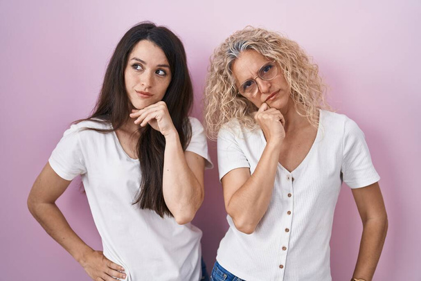 Mother and daughter standing together over pink background with hand on chin thinking about question, pensive expression. smiling with thoughtful face. doubt concept.  - Foto, Imagem