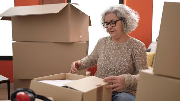 Жінка середнього віку з сірим волоссям розпаковує картонну коробку в новому будинку
 - Фото, зображення