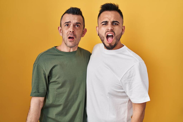 Ομοφυλόφιλο ζευγάρι στέκεται πάνω από κίτρινο φόντο θυμωμένος και τρελός ουρλιάζοντας απογοητευμένοι και έξαλλος, φωνάζοντας με θυμό. οργή και επιθετική αντίληψη.  - Φωτογραφία, εικόνα