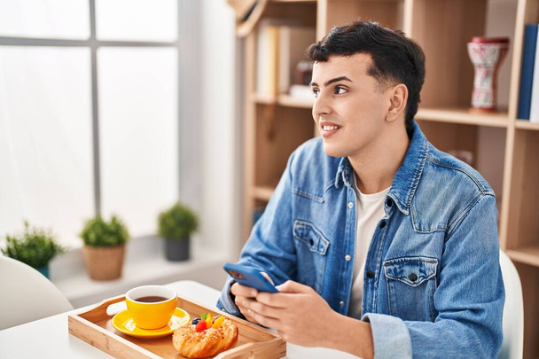 Молодий небінарний чоловік використовує смартфон, сніданок вдома
 - Фото, зображення
