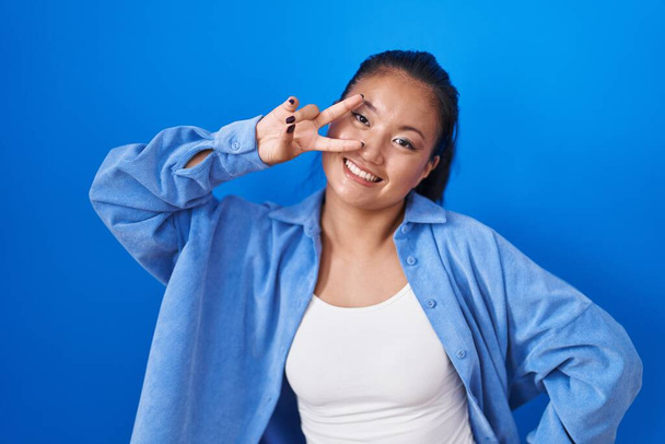 Asiática joven mujer de pie sobre fondo azul haciendo símbolo de la paz con los dedos sobre la cara, sonriendo alegre mostrando victoria  - Foto, Imagen