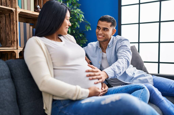 Junges lateinisches Paar erwartet Babybauch auf Sofa zu Hause - Foto, Bild
