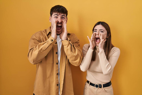 Νεαρό ζευγάρι Ισπανών στέκεται πάνω από κίτρινο φόντο φωνάζοντας θυμωμένος δυνατά με τα χέρια πάνω από το στόμα  - Φωτογραφία, εικόνα