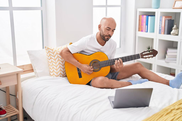 młody łysy człowiek mając online klasę gitara klasyczna siedzi na łóżku w sypialni - Zdjęcie, obraz