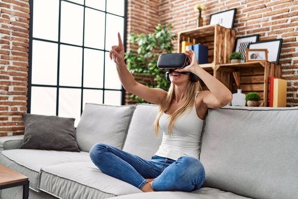 Νεαρή ξανθιά γυναίκα παίζει βιντεοπαιχνίδι χρησιμοποιώντας γυαλιά εικονικής πραγματικότητας στο σπίτι - Φωτογραφία, εικόνα
