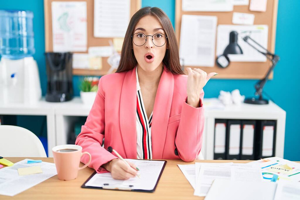Jonge Spaanse vrouw werkt op kantoor draagt een bril verrast wijzen met de hand vinger naar de zijkant, open mond verbaasd uitdrukking.  - Foto, afbeelding