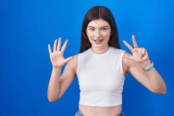 Nuori valkoihoinen nainen seisoo sinisen taustan yläpuolella osoittaen sormilla numero kahdeksan hymyillen itsevarmana ja onnellisena.  - Valokuva, kuva