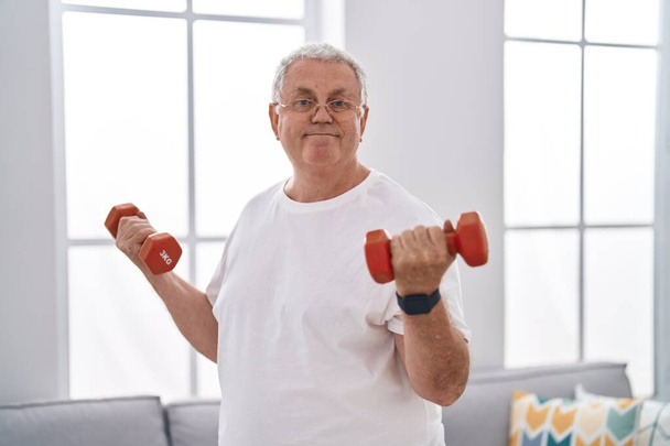 Седовласый мужчина средних лет уверенно улыбается, используя гантели для тренировки дома - Фото, изображение