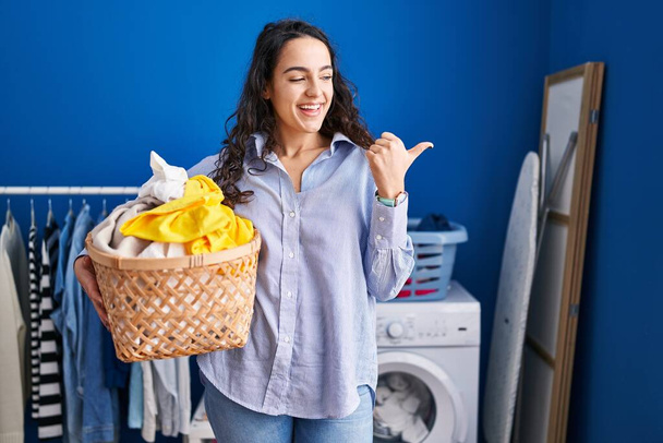 Young brunette woman holding laundry basket pointing thumb up to the side smiling happy with open mouth  - Foto, Imagem